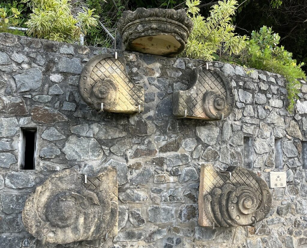 Cinco peças de pedra lavrada fixadas num muro na Fortaleza do Morro de São Paulo: uma coroa e quatro volutas, possivelmente do frontão da capela primitiva de Nossa Senhora da Luz